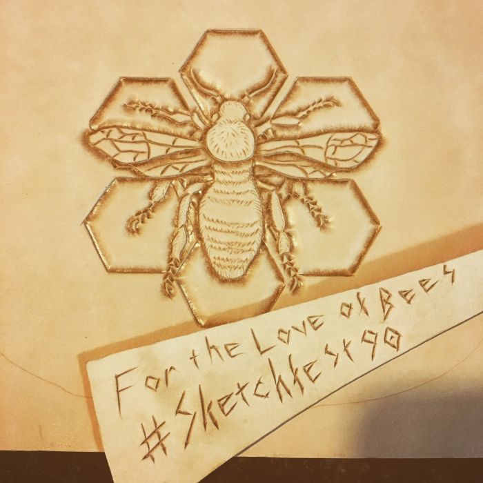 A bee has been tooled on a piece of undyed leather; the bee is depicted against six hexagons in a honeycomb pattern. Another piece of leather lays across this piece, with For the Love of Bees #Sketchfest 90 scratched into it.